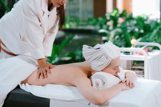 Beautiful young woman received a back massage on a spa bed from masseuse. Attractive female relaxes deeply by skilled hands of the massage therapist. Surrounded with nature. Side view. Tranquility.