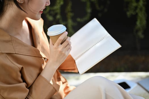 Peaceful young woman drinking coffee from paper cup and reading book outdoor.