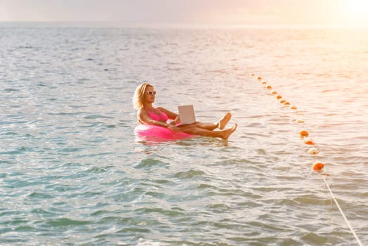 Woman works on laptop in sea. Freelancer, blond woman in sunglases floating on an inflatable big pink donut with a laptop in the sea at sunset. People summer vacation rest lifestyle concept