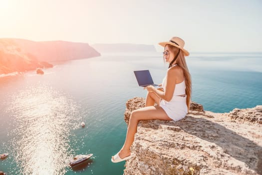 Successful business woman in yellow hat working on laptop by the sea. Pretty lady typing on computer at summer day outdoors. Freelance, travel and holidays concept.