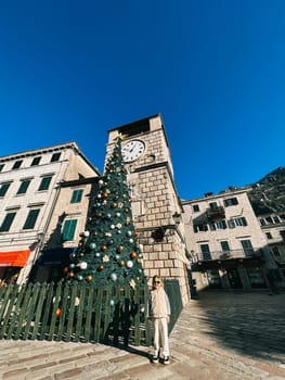 Little girl stands near a Christmas tree in front of an ancient building with a clock. High quality photo