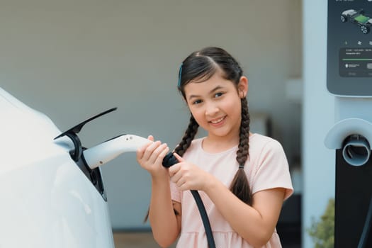 Happy little young girl learn about eco-friendly and energy sustainability as she recharge electric vehicle from home EV charging station. EV car and sustainable future generation concept. Synchronos