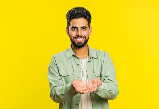 Free gifts and presents for you. Indian man opening hands, excitement, preparing surprise, sharing love emotions, congratulating, charity donation. Arabian guy isolated on yellow studio background