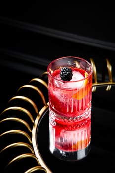 red cocktail in a transparent glass with round ice, whipped cream and black berry on top