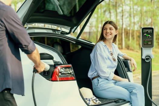 Lovely young couple recharging battery for electric car while relaxing and sitting on the trunk during road trip travel EV car in autumnal forest. Eco friendly travel on vacation during autumn. Exalt