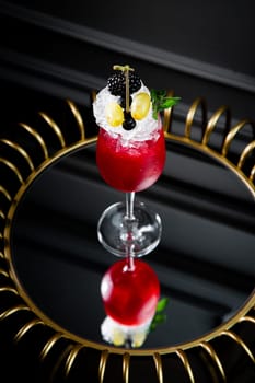 red cocktail in a transparent glass with round ice, whipped cream and black berry on top