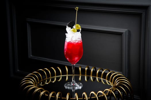 red cocktail in a transparent glass with round ice, whipped cream and black berry on top