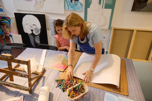 Beautiful blonde female painter artist teaching paint art to a little student girl in creative workshop. Visual and fine art lesson in cozy studio. People. Creativity. Inspiration.