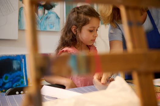 View though a blurred wooden easel of a cute little kid girl learning painting and art in creative workshop or art gallery, with painted pictures exhibited on a white wall background. People. Fine art