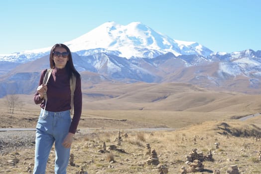 A female traveler with a backpack on her back admires the snow-covered Elbrus on a clear autumn day. View of Elbrus, North Caucasus, Russia.