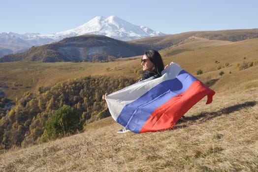 A young brunette woman sits on a stone and looks at the Caucasus Mountains around, a Russian flag flutters in her hands. Tricolor against the backdrop of snow-capped Mount Elbrus.