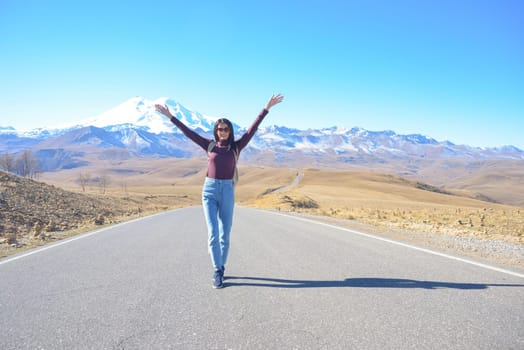 A happy female traveler with a backpack behind her back and her arms raised up rejoices at her trip to the mountains. Elbrus on a clear autumn day. View of Elbrus, North Caucasus, Russia.