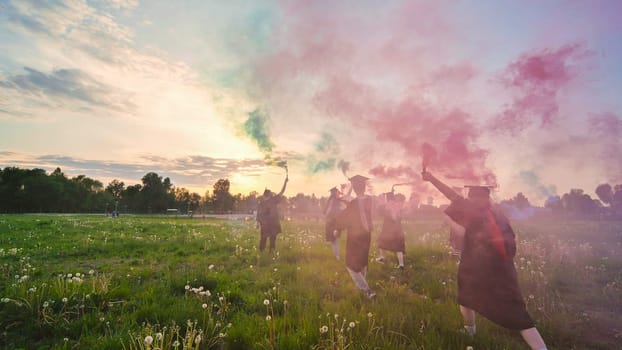 Graduates in costume walk with a smoky multi-colored smoke at sunset.