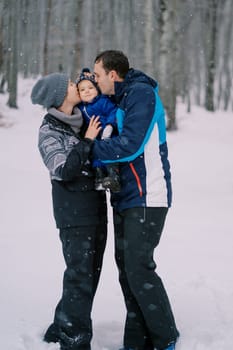 Mom and dad kiss a small child in their arms while standing in a snowy forest. High quality photo