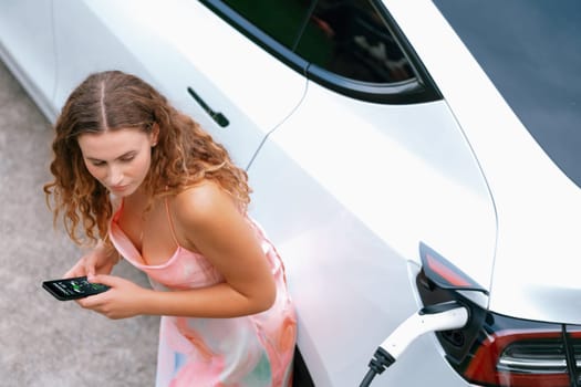 Aerial top view eco-friendly woman recharge electric vehicle from EV charging station, using EV technology utilization for tracking energy usage to optimize battery charging on smartphone. Synchronos