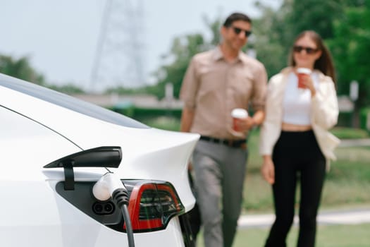 Young couple with coffee cup, recharge electric car's battery from EV charging station in green city park. Sustainable and eco friendly EV car with urban and shopping lifestyle. Expedient