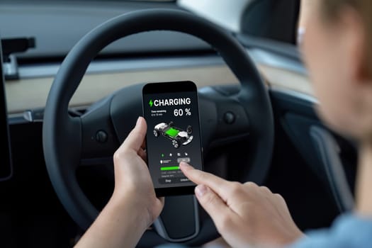 Holiday vacation road trip with environmental-friendly car concept. Eco-conscious woman on driver seat checking EV car's battery status display on smartphone during car travel. Perpetual