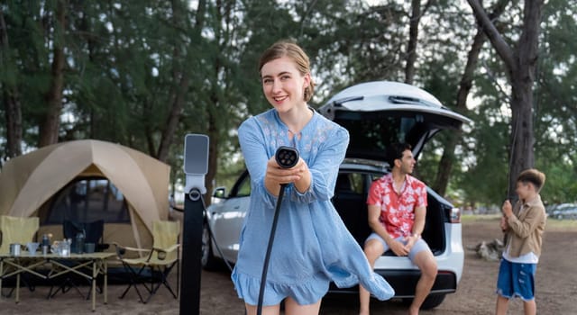 Outdoor adventure and family vacation camping at sea travel by eco friendly car. Cheerful woman or mother holding, pointing EV charger point with playful and happiness posture in campsite. Perpetual