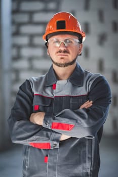 Vertical portrait of a Caucasian bearded worker in overalls, an orange hard hat and safety glasses.