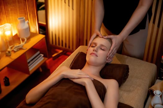 Caucasian woman enjoying relaxing anti-stress head massage and pampering facial beauty skin recreation leisure in warm candle lighting ambient salon spa in luxury resort or hotel. Quiescent