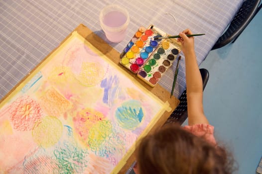 Top view of little child girl painter artist sitting at desk with painting accessories, holding paintbrush, mixing colors on palette, drawing on white paper sheet using a watercolor painting technique