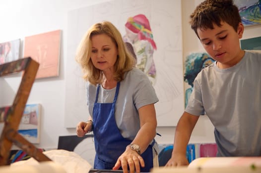 Adorable Caucasian teenage school boy learning painting and art, standing with a female paint teacher artist by a desk, working together in creative visual art workshop