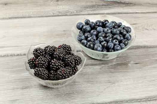 Two small glass bowls, one with blackberries, other blueberries, on gray wood desk.
