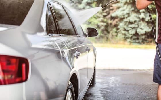 Man washing his silver car at self serve carwash, water spraying from high pressure jet nozzle.