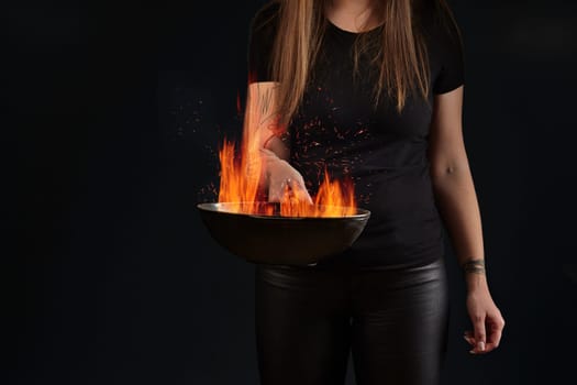 Young brunette girl with tattooed hands and long hair, dressed in leather leggings and t-shirt. Holding a wok pan with fire against black studio background. Cooking concept. Close up, copy space