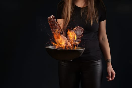 Brunette female with tattooed hands, dressed in leggings and t-shirt. Holding a wok pan with fire and frying two beef steaks against black studio background. Cooking concept. Close up, copy space