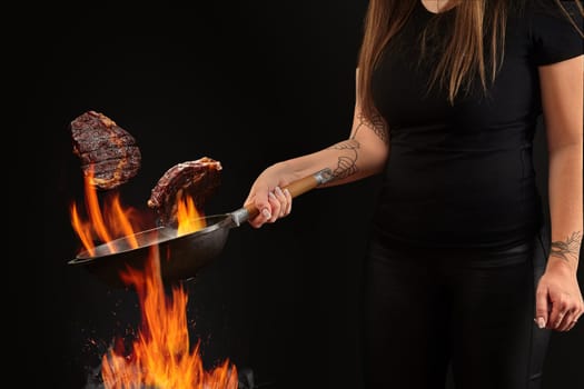 Brunette cook with tattooed hands, dressed in leggings and t-shirt. Holding wok pan above fire and frying two beef steaks against black background. Cooking concept. Close up, copy space, side view