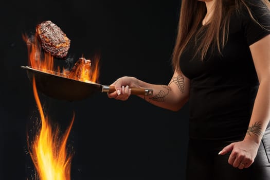 Woman with tattooed hands, dressed in leggings and t-shirt. Holding burning wok pan above fire and frying two beef steaks against black background. Cooking concept. Close up, copy space, side view