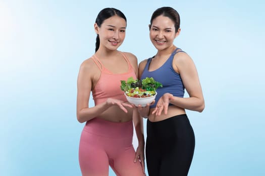 Two young sporty Asian women in sportswear holding salad bowl fill with fruit and vegetable. Natural youthful and fit body lifestyle people with balance nutrition on isolated background. Vigorous