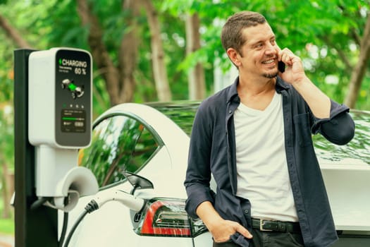 Man talking on smartphone while recharging electric car battery charging from EV charging station during vacation holiday road trip at national park or summer forest. Exalt