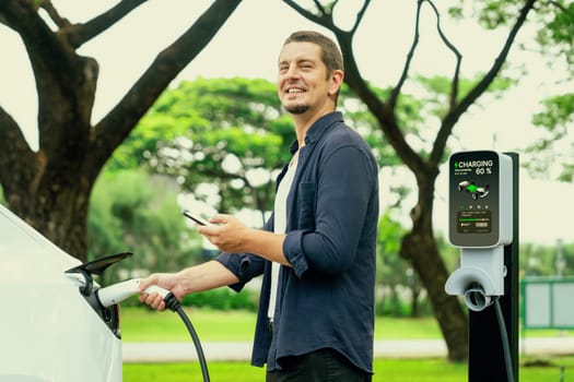 Man using smartphone online banking application to pay for electric car battery charging from EV charging station during vacation holiday road trip at national park or summer forest. Exalt