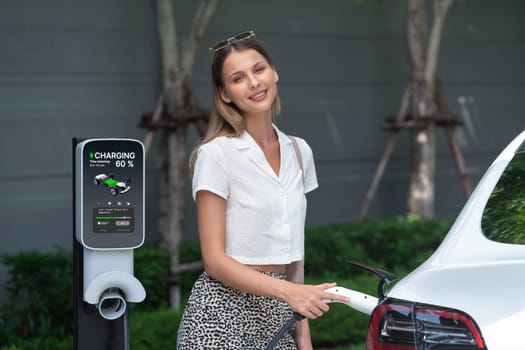 Young woman travel with EV electric car charging in green sustainable city outdoor garden in summer. Urban sustainability lifestyle by green clean rechargeable energy of electric BEV vehicle innards