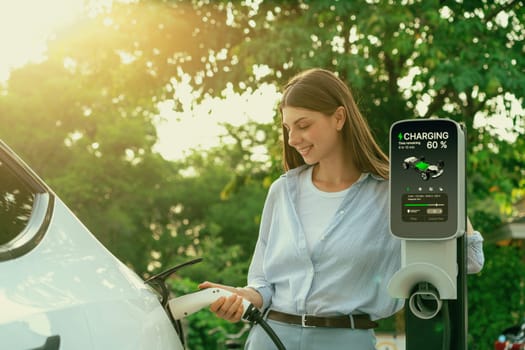 Young woman recharging battery for electric car during road trip travel EV car in natural forest or national park with sunrise lighting. Eco friendly travel during vacation and holiday. Exalt