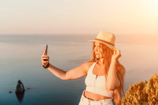 Selfie woman in a hat, white tank top, and shorts captures a selfie shot with her mobile phone against the backdrop of a serene beach and blue sea