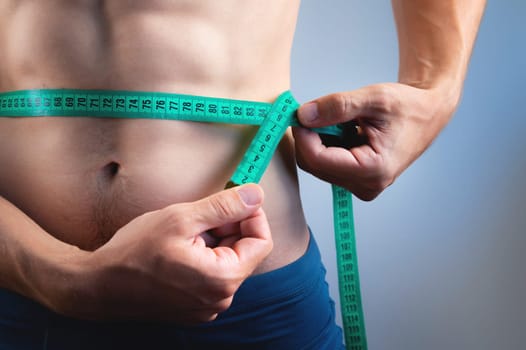 A muscular athlete poses shirtless and measures his waistline with a measuring tape. Close-up of an athlete measuring his body volume and the result of diet and training