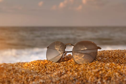 Side view of sunglasses on the beach near the turquoise sea with sand. Summer, beach holiday, stylish sunglasses, no people.