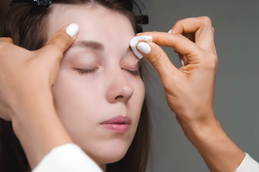 In a beauty salon, a master cosmetologist applies makeup and wipes eyebrows with a cotton pad. Close-up of the model's face during the procedure.