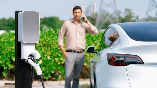 Man talking on the phone while recharge EV car battery at charging station connected to power grid tower electrical as electrical industry for eco friendly car utilization. Panorama Expedient