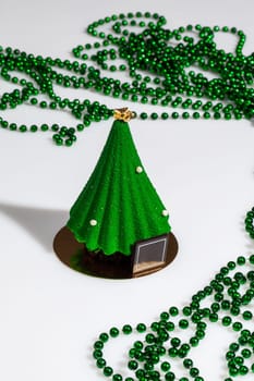 Christmas tree shaped pastry covered with scabrous green icing with golden topper and sugar drops on white background with strings of green beads. Festive decor and treats. Handmade authors dessert