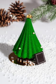 Green Christmas tree shaped cake decorated with edible gold on top, chocolate and sugar beads on white surface with fir branch and cones. Creating festive atmosphere with authors handmade dessert