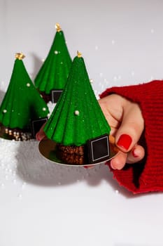 Female hand holding out Christmas tree shaped pastry covered with scabrous green icing with golden topper and sugar drops on serving cardboard. Festive treats concept. Handmade authors dessert