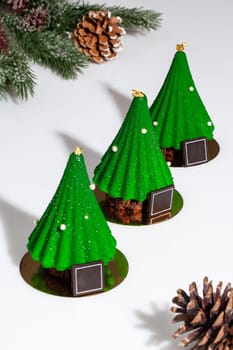 Pistachio mousse, strawberry marmalade and chocolate brownie cakes in form of Christmas trees covered with green icing, decorated with edible gold and sugar drops on table with cones. Festive desserts