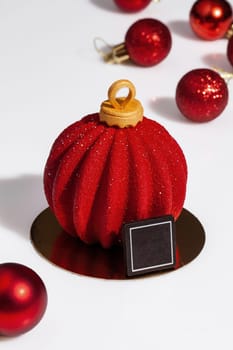 Red sparkling Christmas tree toys and ball shaped cake on white surface. Festive handmade dessert of chocolate mousse, cherry compote, hazelnut and almond biscuit covered with colored sugar icing