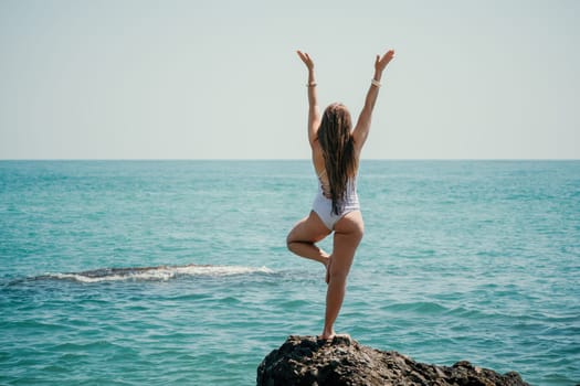 Woman sea yoga. Back view of free calm happy satisfied woman with long hair standing on top rock with yoga position against of sky by the sea. Healthy lifestyle outdoors in nature, fitness concept