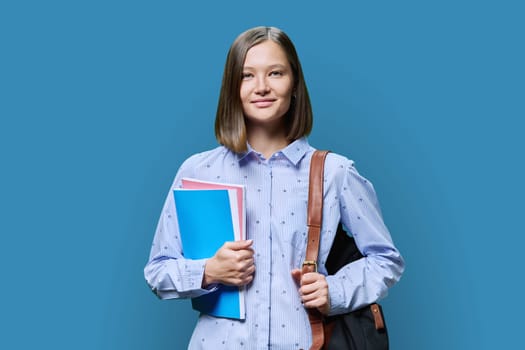 Portrait of young university student with backpack of textbooks on blue studio background. Smiling confident positive 20s female looking at camera. Education, knowledge, lifestyle, youth concept