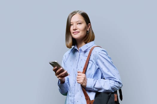 Young woman student with smartphone backpack on grey studio background. Smiling attractive female looking at camera. Using mobile applications apps for work business study leisure, technology, people